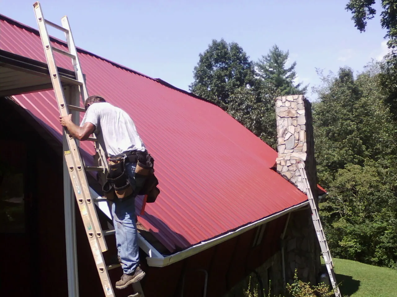 Roofer on a ladder beside a red metal roof and stone chimney — Soffit & Fascia Repair by Roofing Brothers in Kountze